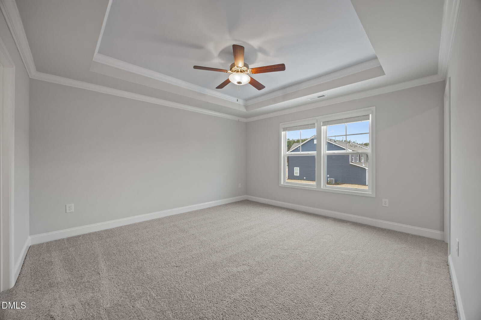 A spacious, empty bedroom with light gray walls, a tray ceiling with a ceiling fan, two large windows letting in natural light, and light-colored carpet flooring.