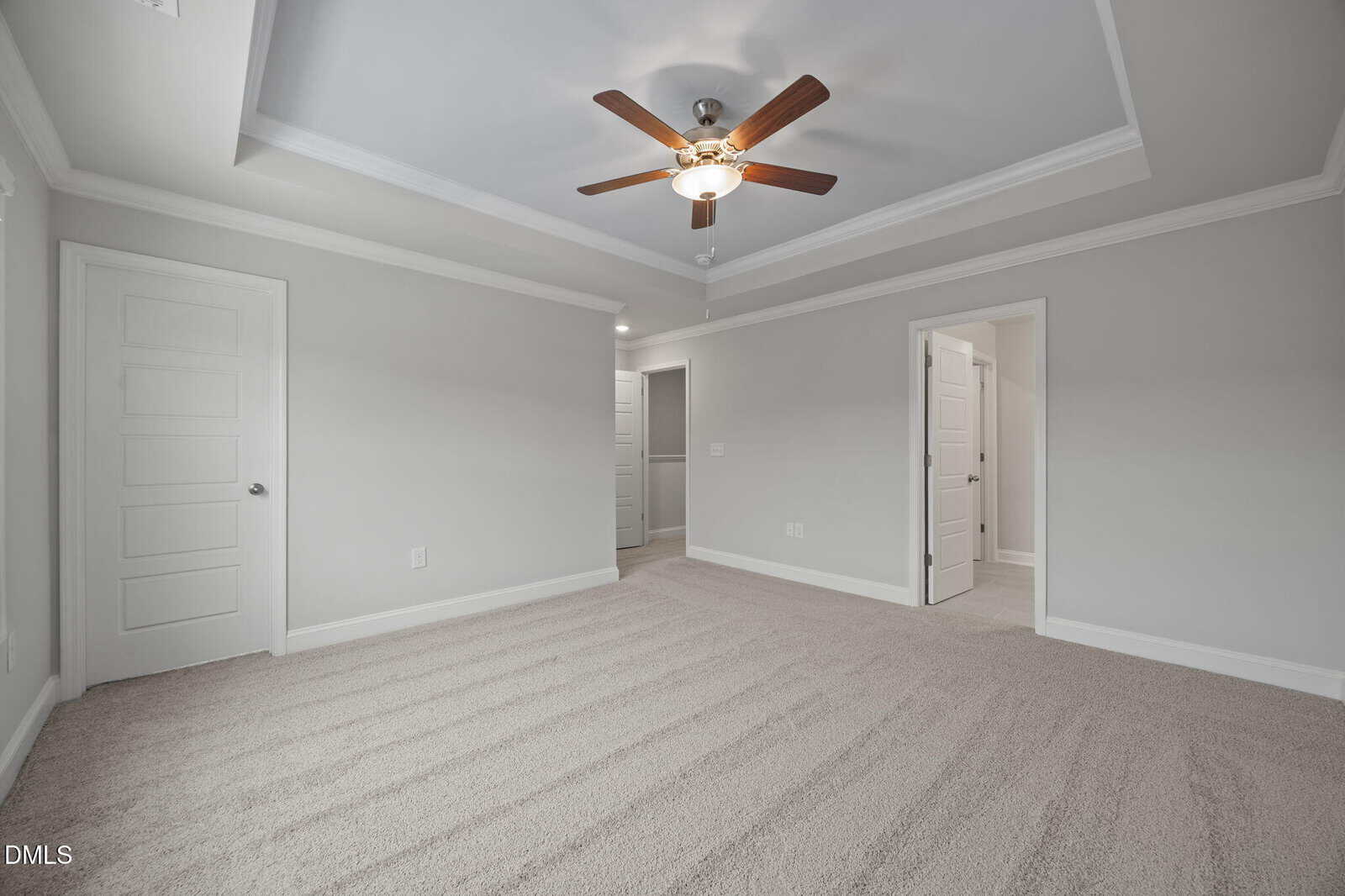 Empty bedroom with light gray walls, beige carpet, a ceiling fan with light, tray ceiling, and three white doors leading to other rooms or closets. The room is well-lit with natural and recessed lighting.