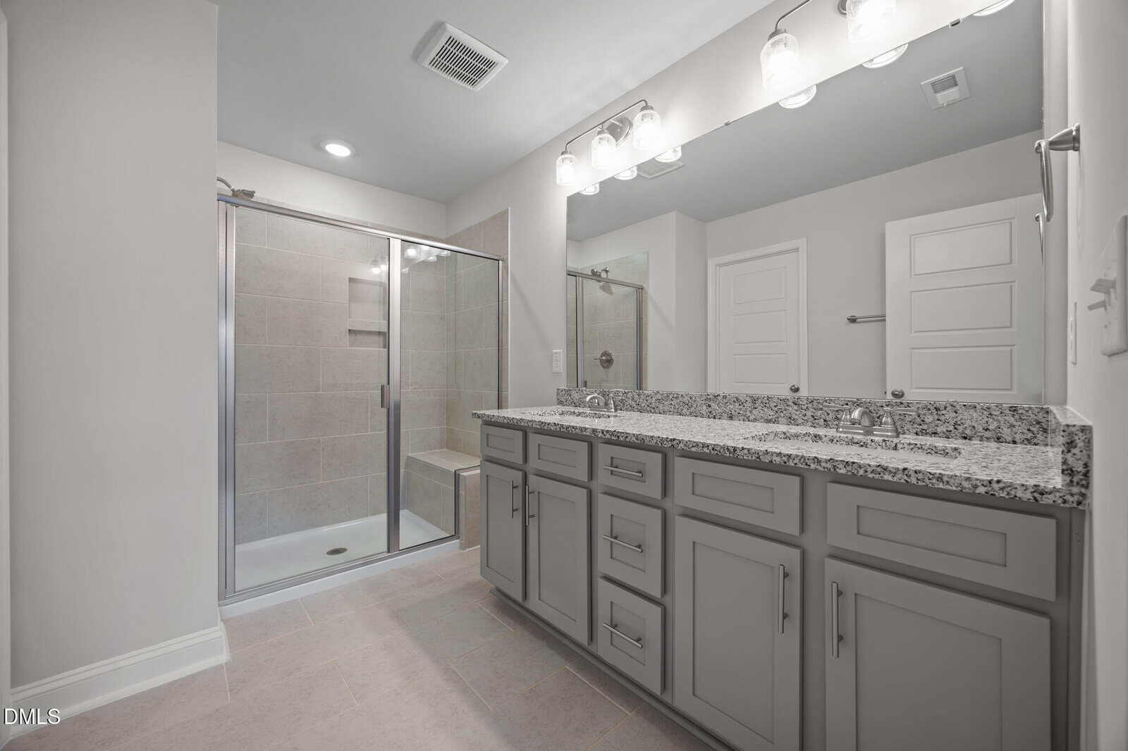 Modern bathroom with a granite double-sink vanity, gray cabinets, a large mirror with lights, and a glass-enclosed shower featuring a tiled interior and built-in bench. Walls and doors are white.