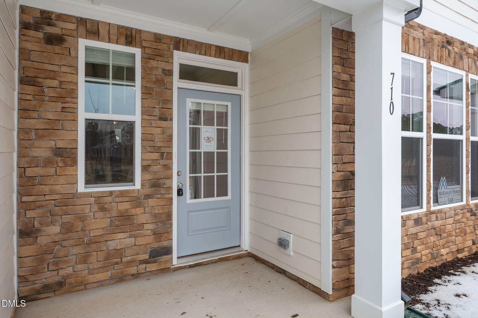 A house entrance with a light blue front door, two tall windows, a white column marked "710," and exterior walls featuring tan siding and brown stone accents.