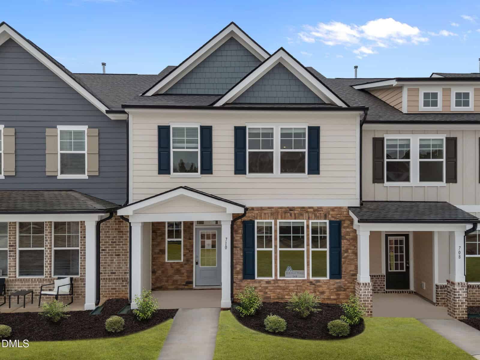 A two-story townhouse with a brick and siding exterior, blue shutters, a covered entryway, and neatly landscaped front yard, set against a bright blue sky with scattered clouds.