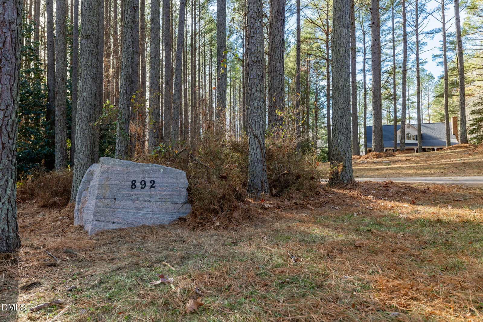 A large rock with the house number 892 etched on it sits among tall pine trees near a driveway; a house is partially visible in the background.