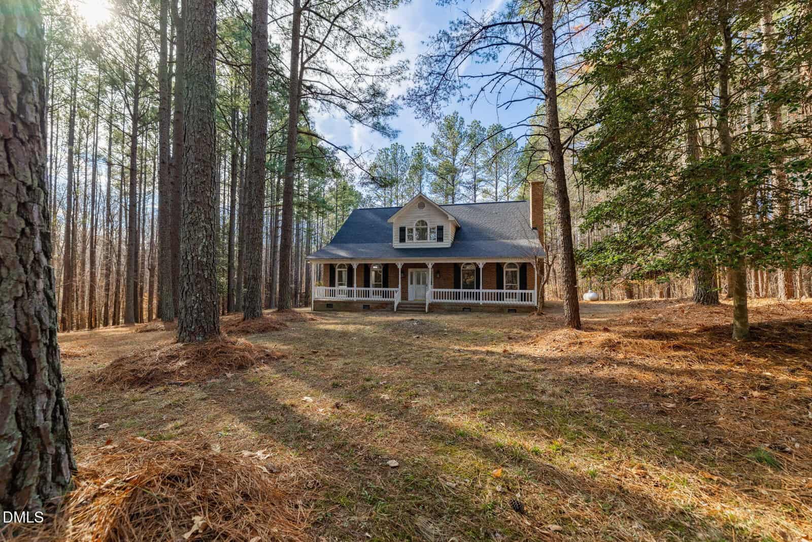 A two-story house with a covered front porch sits in a wooded area surrounded by tall pine trees, with sunlight filtering through the branches and pine needles covering the ground.