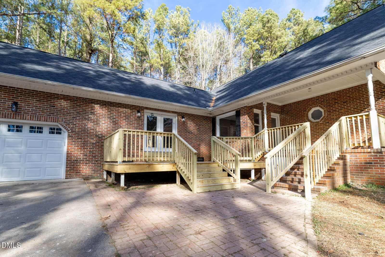 A brick house with a black roof features two sets of wooden steps leading to a porch and a white door; a garage door is visible on the left, with trees and blue sky in the background.