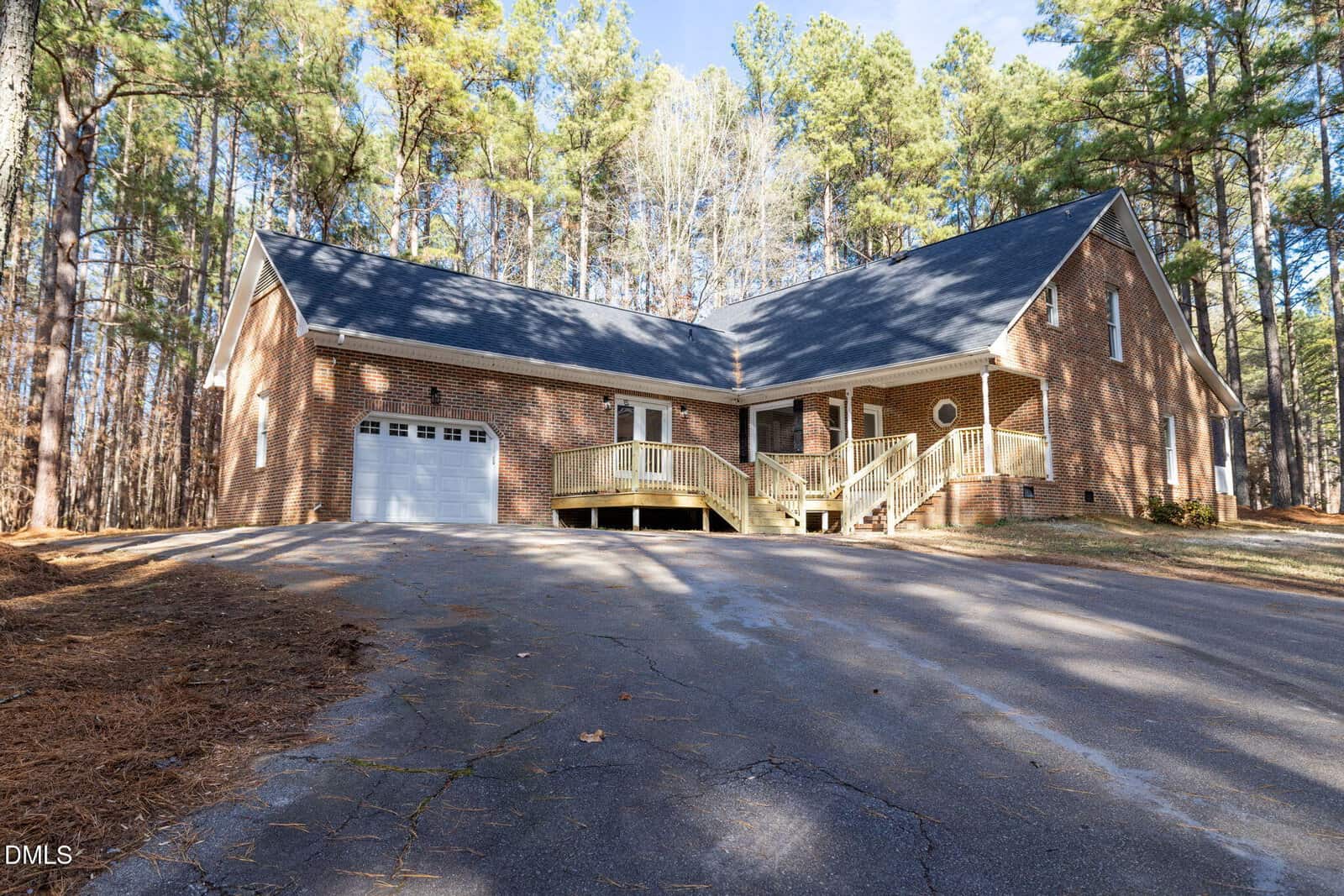 A brick house with a dark shingle roof sits among tall pine trees. It has a single-car garage, a front porch with steps, and a paved driveway leading up to the house. The yard is covered with pine needles.