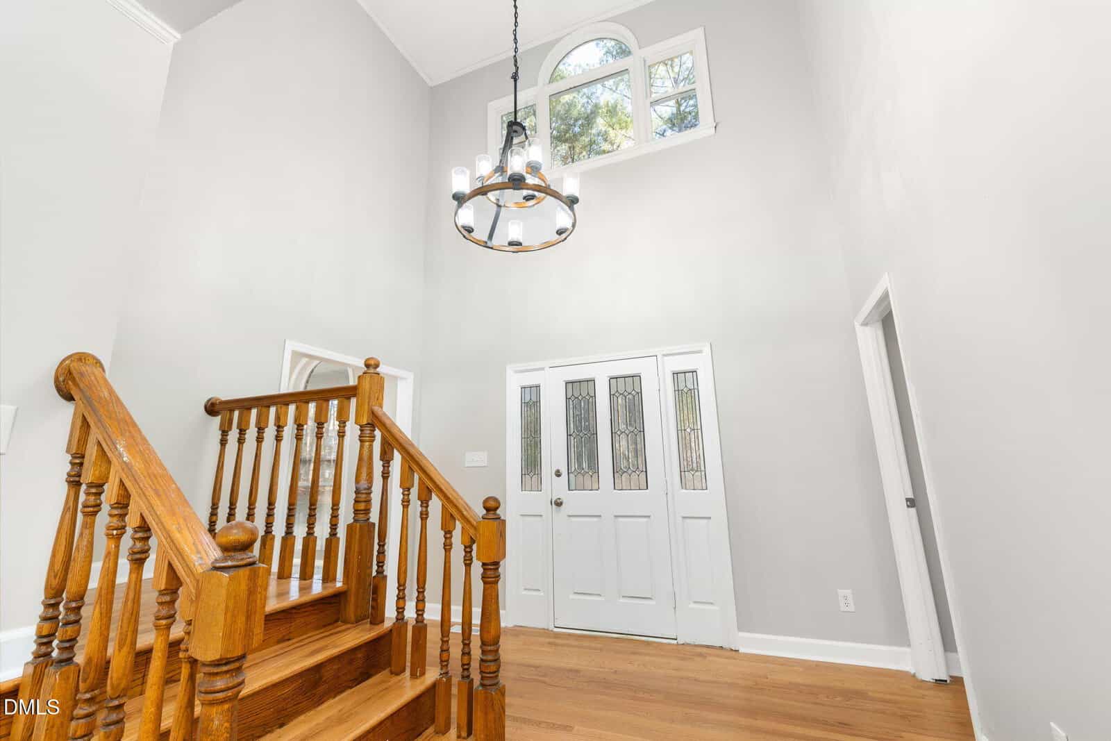 A bright entryway with a white double front door featuring glass panels, a wooden staircase with spindles, a circular chandelier, hardwood floors, and high ceilings with a window above the door.