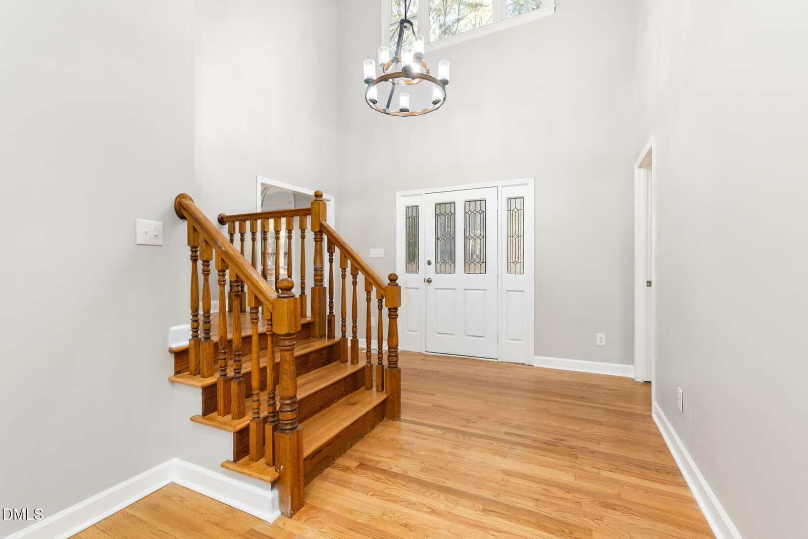 Entryway with hardwood floors, white walls, wooden staircase with banister, double glass-paneled front doors, chandelier, and natural light from a high window above the doors.