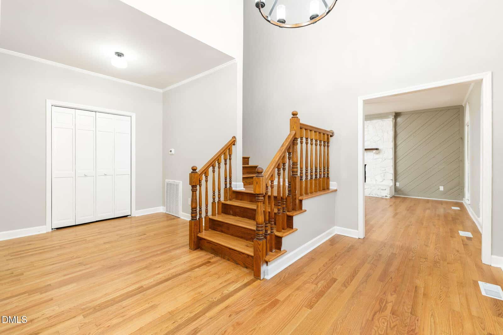 A bright interior with light wood floors, wooden stairs with spindles, white walls, closets with double doors, and an open doorway to another room with a fireplace and accent wall.