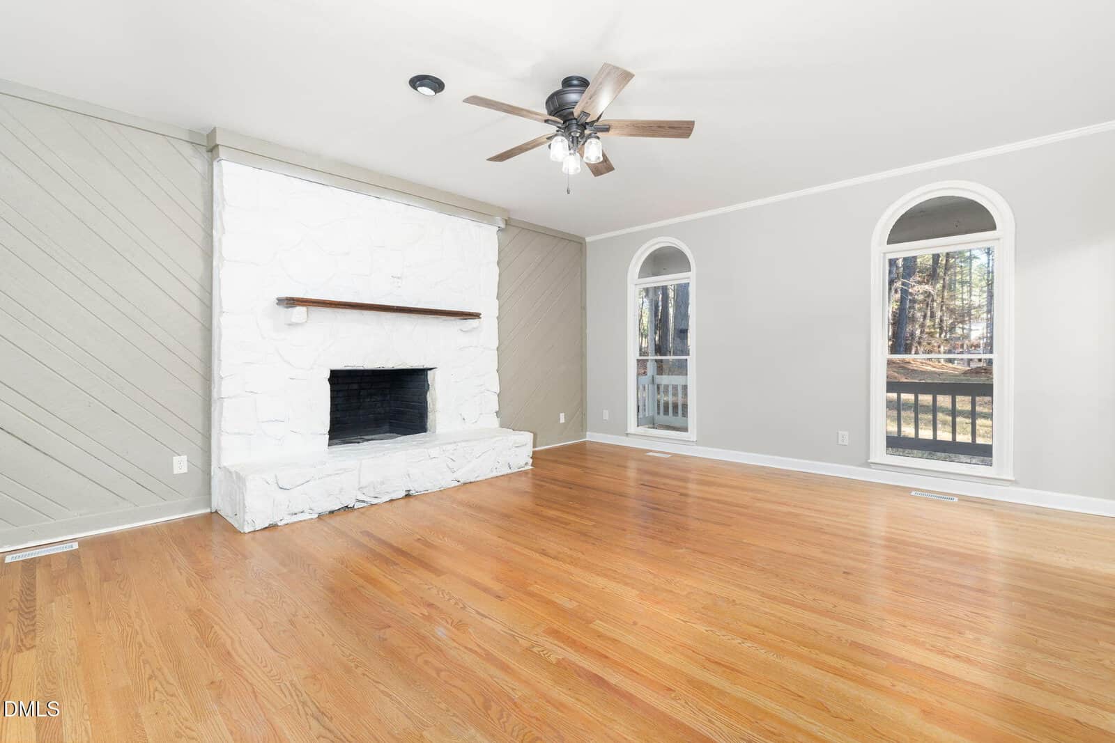 Bright living room with hardwood floors, white stone fireplace, ceiling fan, and two tall arched windows letting in natural light. Room has light gray walls and a simple, open layout.