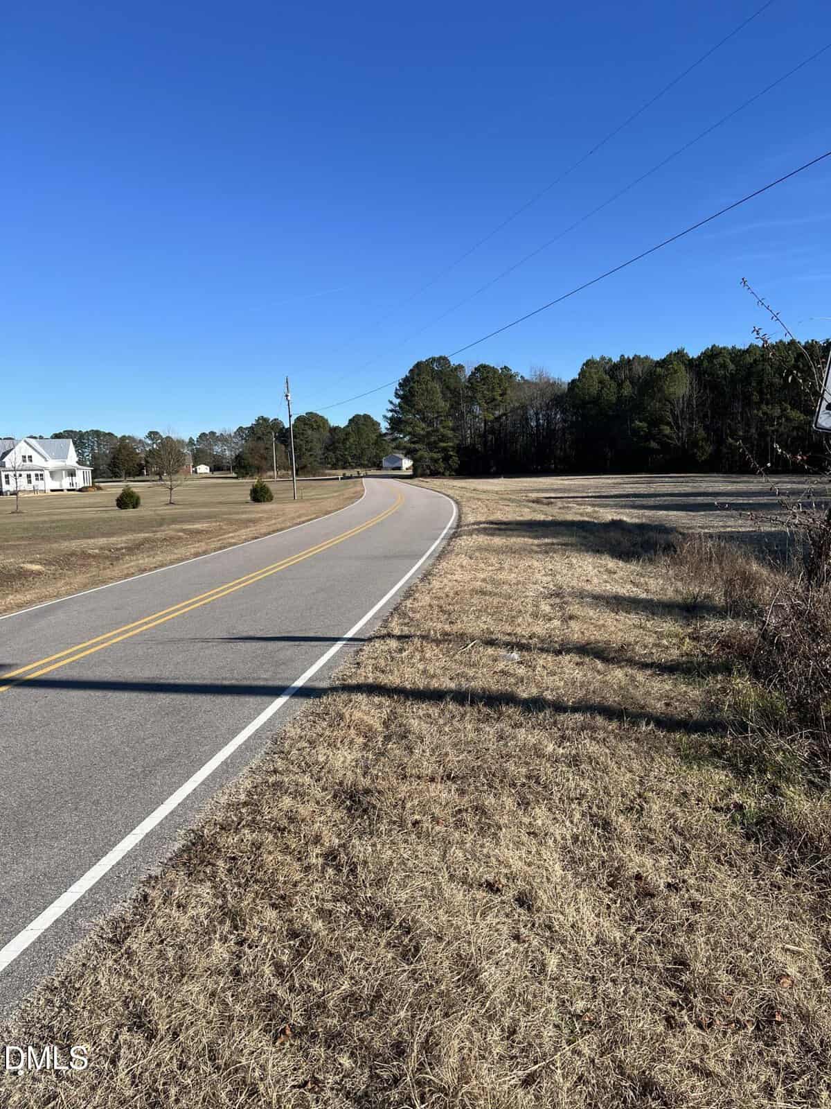 A rural two-lane road curves through dry grassland under a clear blue sky, with a few scattered houses and trees in the background. Power lines run alongside the road.