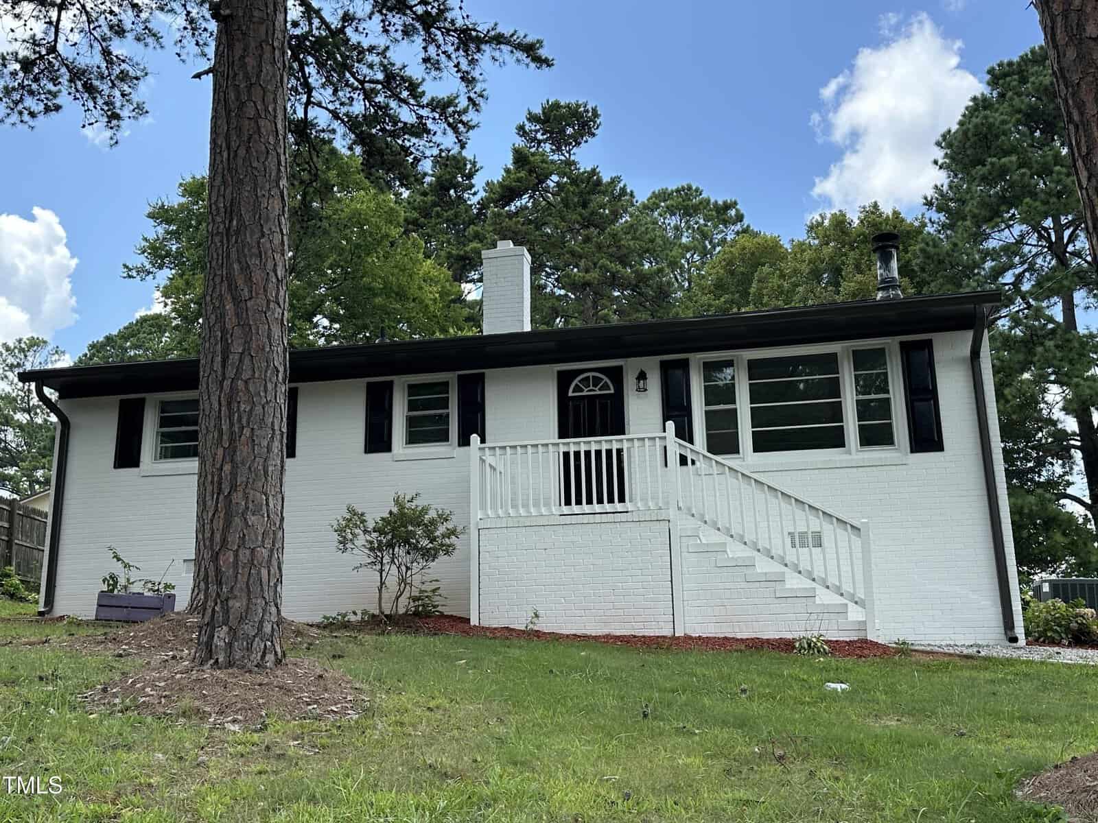 A single-story white brick house with black shutters at 1220 Roanoke Avenue, Henderson, features a central front door, white railing and steps, all surrounded by grass, trees, and a blue sky with clouds.