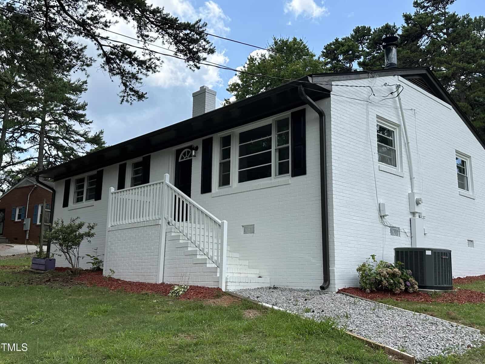 Single-story white brick house with black shutters and trim, front stoop with white railing, gravel walkway, small shrubs, central air unit, and trees in the background under a partly cloudy sky.