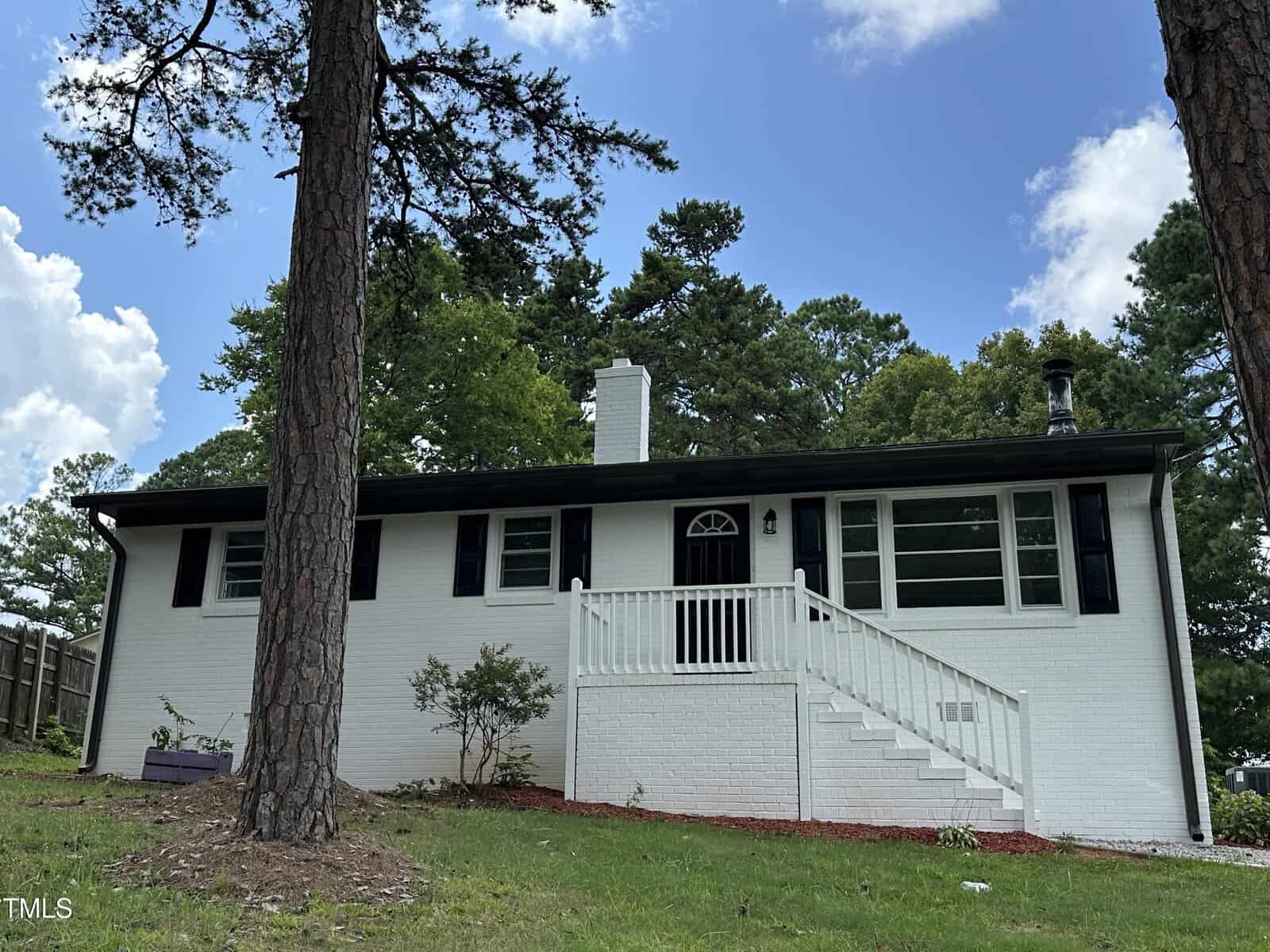 A white brick, single-story house with black shutters and trim, a central chimney, and a front porch with white railings. Stairs lead up to the porch, and tall pine trees are in the yard. The sky is partly cloudy.
