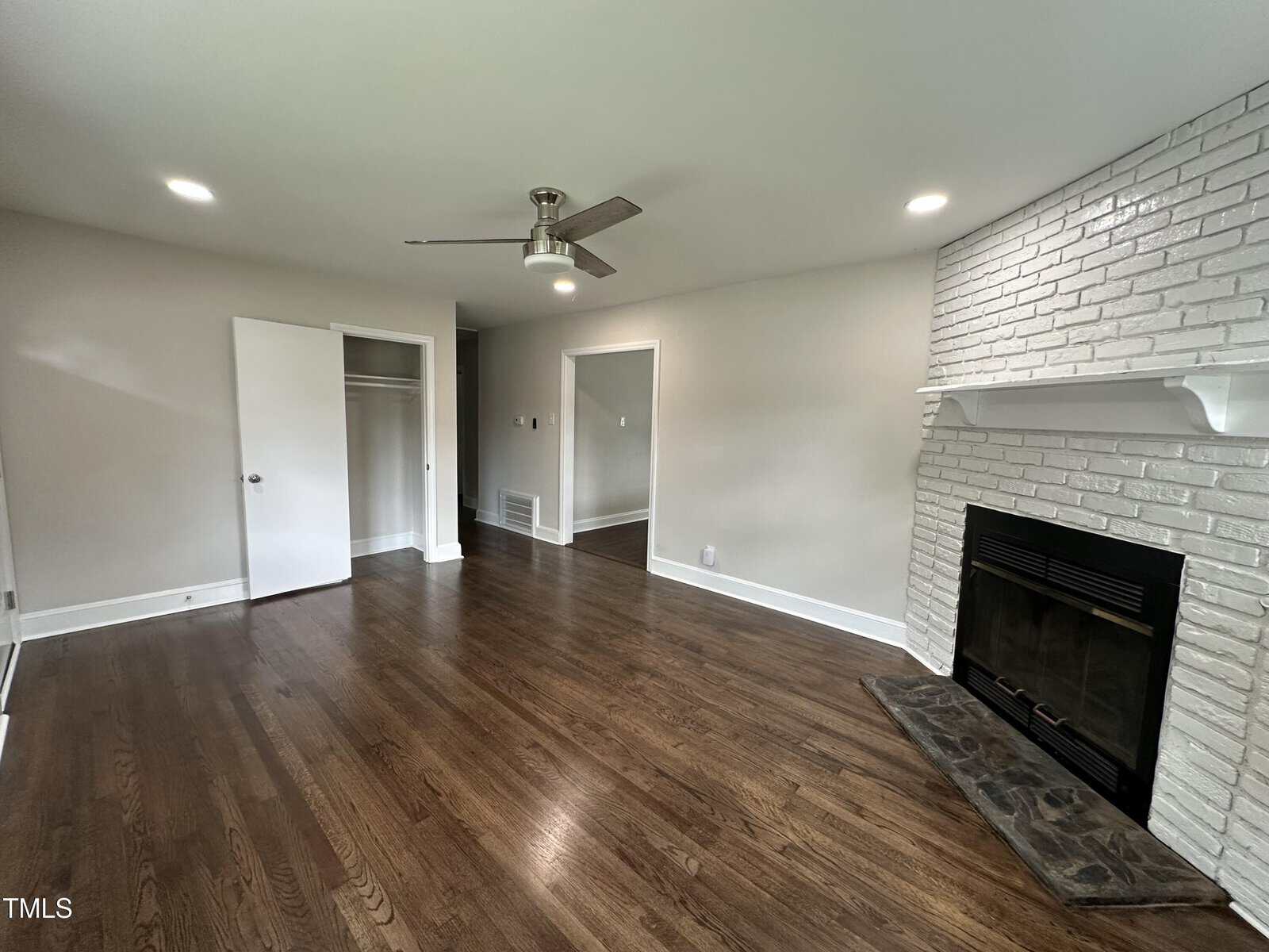 A room with hardwood floors, white walls, recessed ceiling lights, a ceiling fan, an open closet, and a white painted brick fireplace with a mantle. The room is empty and well-lit by natural and artificial light.