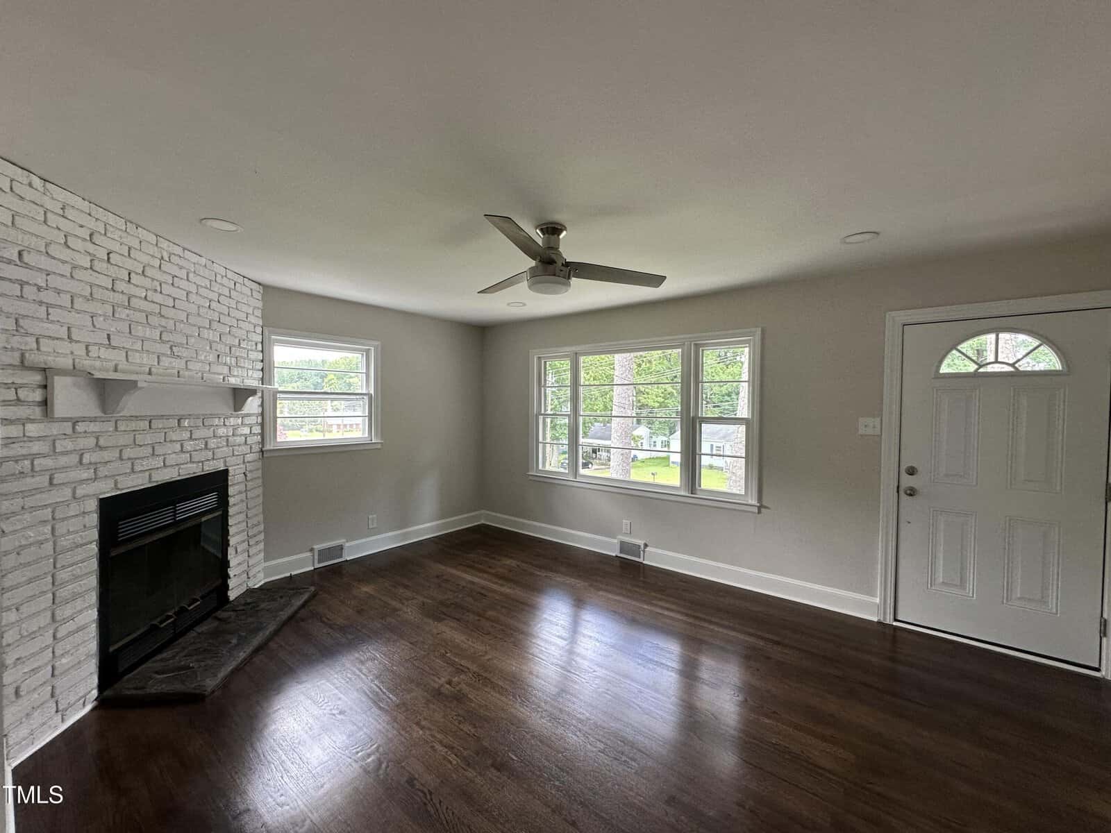 A living room with dark wood floors, a white brick fireplace, light gray walls, three windows, a ceiling fan, and a white door with a window. The room is empty and receives natural light.
