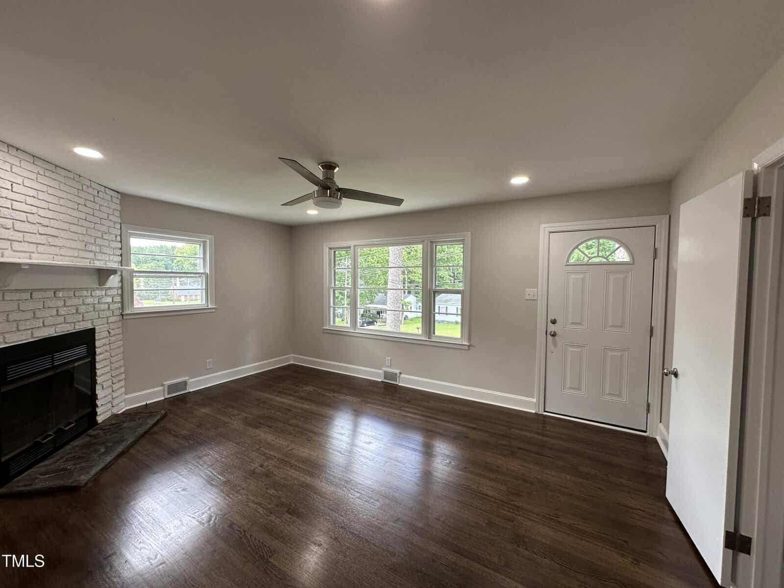 A bright, empty living room with dark wood floors, white brick fireplace, ceiling fan, large windows, and a white front door. The walls are light-colored and natural light fills the room.