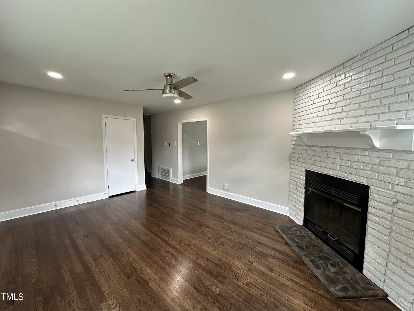 A living room with hardwood floors, a white brick fireplace with a white mantel, a ceiling fan with light, neutral-colored walls, and recessed lighting. There are doorways leading to other rooms.