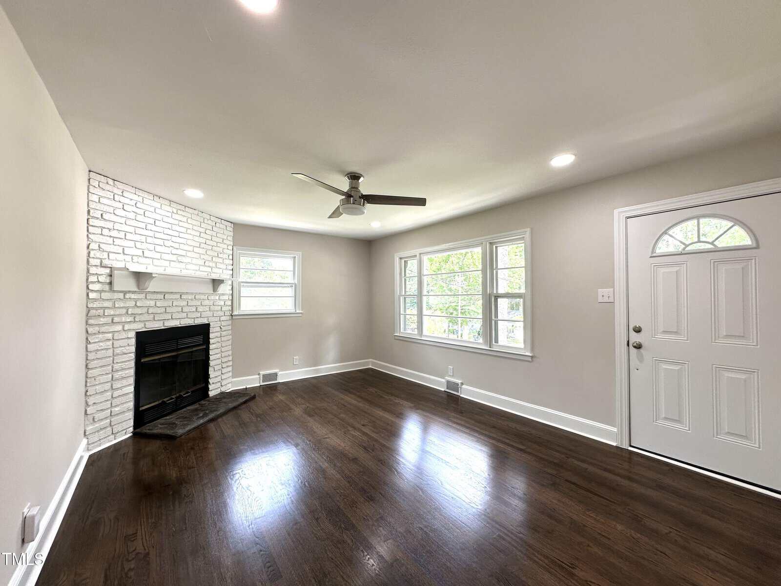 A bright living room with dark wood floors, white walls, a white brick fireplace with a mantle, a ceiling fan, several windows, and a white front door with a semi-circular window at the top.