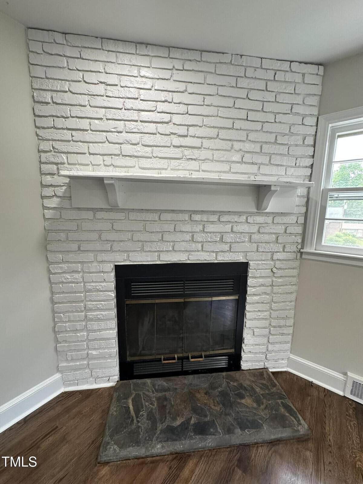 A white-painted brick fireplace with a black metal insert and glass doors, topped by a simple white mantel shelf. The hearth is a slab of dark stone, and a window is visible to the right.