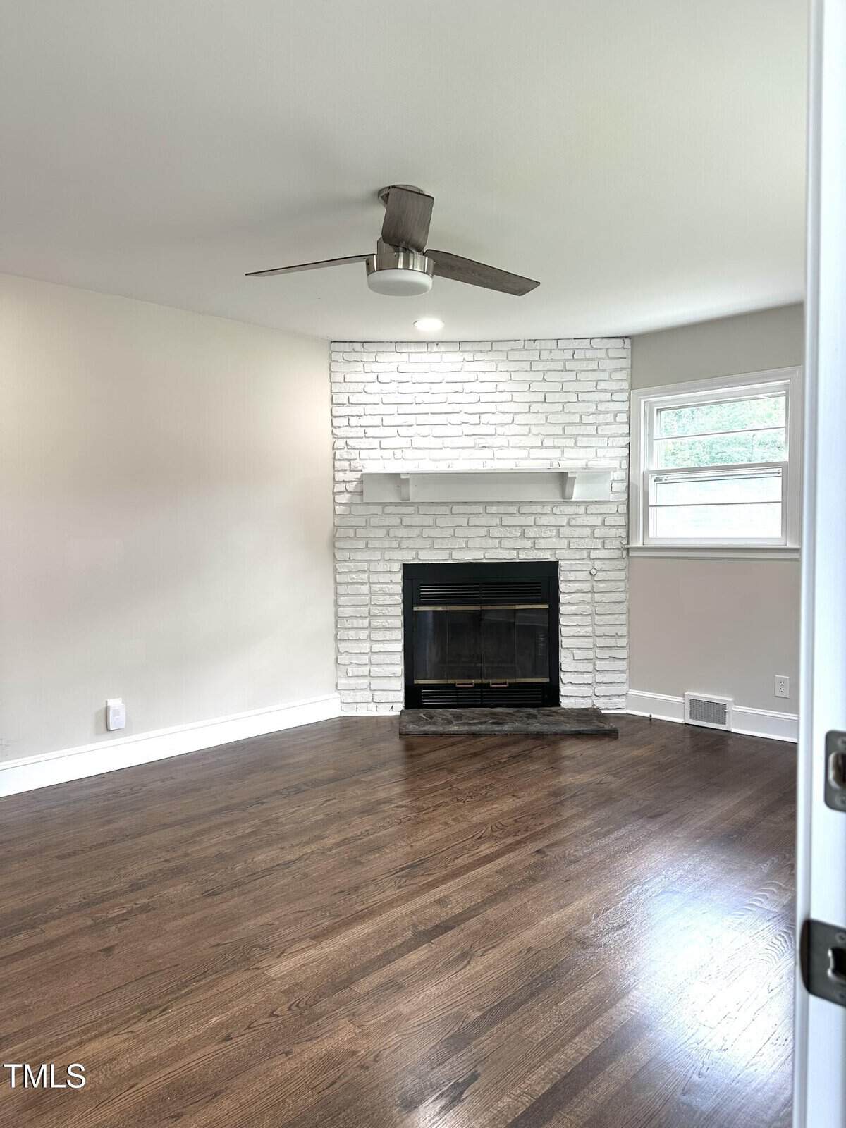A room with dark hardwood floors, beige walls, a white painted brick fireplace with a mantle, a metal ceiling fan, and two windows letting in natural light.