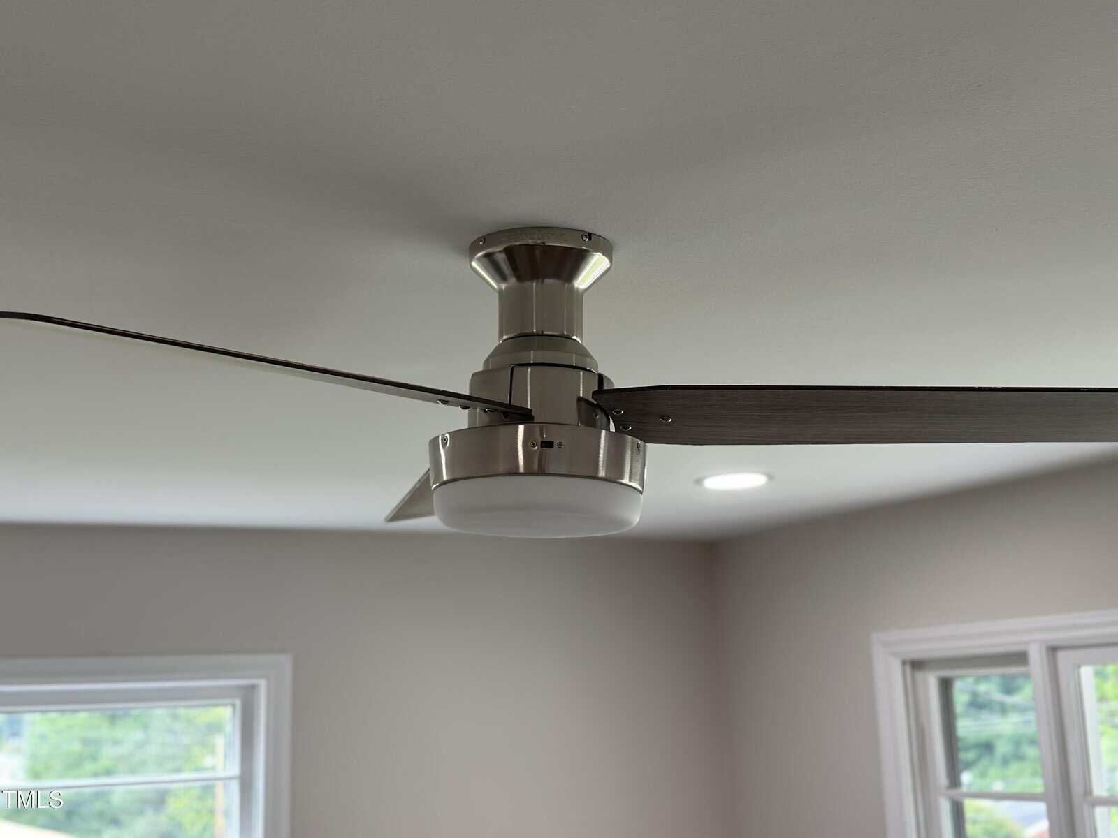Modern ceiling fan with silver base and wooden blades, mounted on a white ceiling. Two windows and recessed lighting are visible in the background.