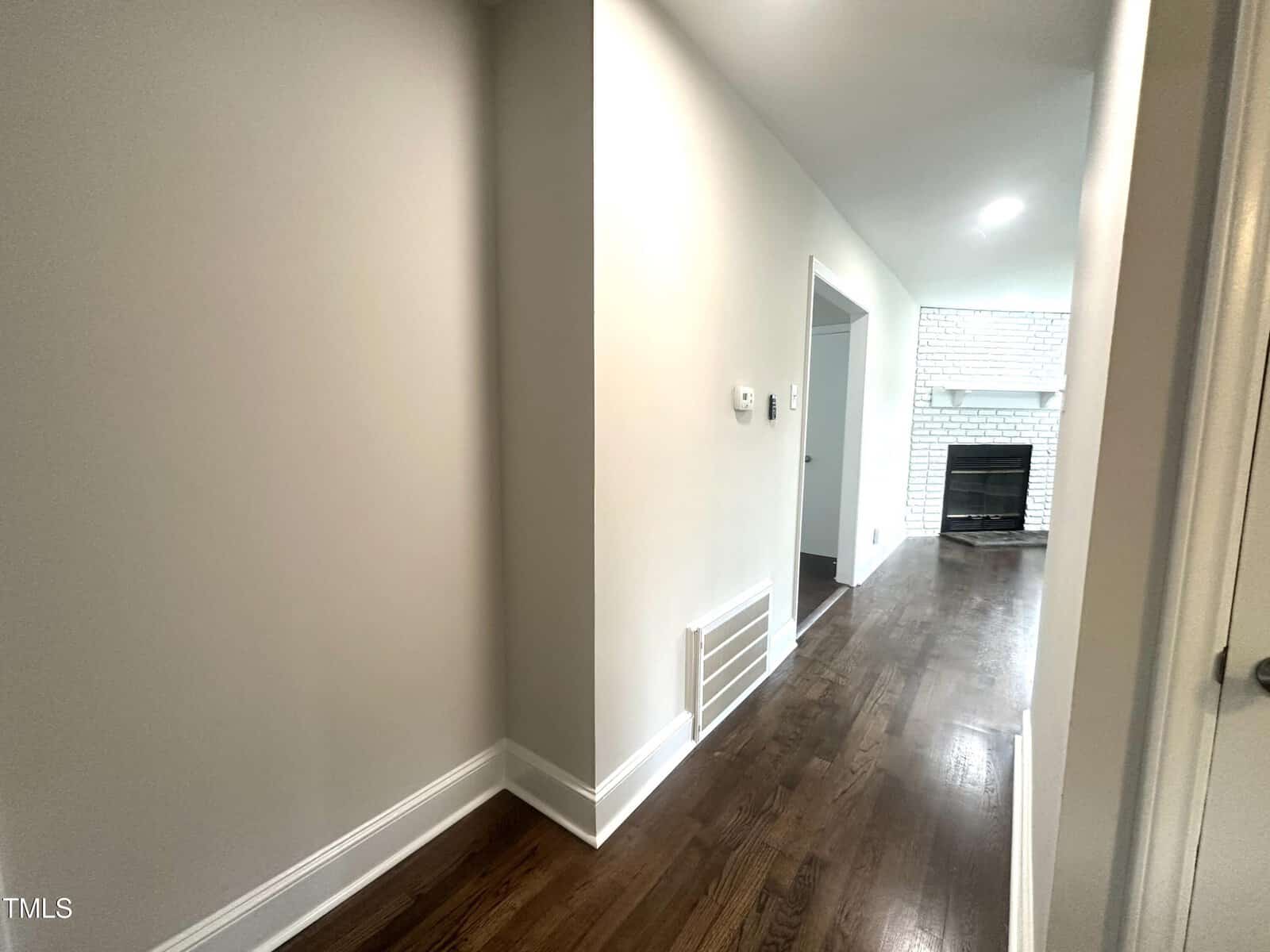 Bright hallway with light-colored walls and dark wood flooring, leading to a living area with a white brick fireplace. Several doorways and a vent are visible along the hallway.