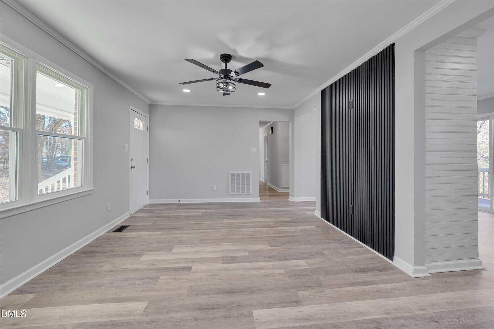 A spacious, empty room with light wood floors, gray walls, large windows, a ceiling fan with light, and a black slatted accent wall beside an open entryway to another space.