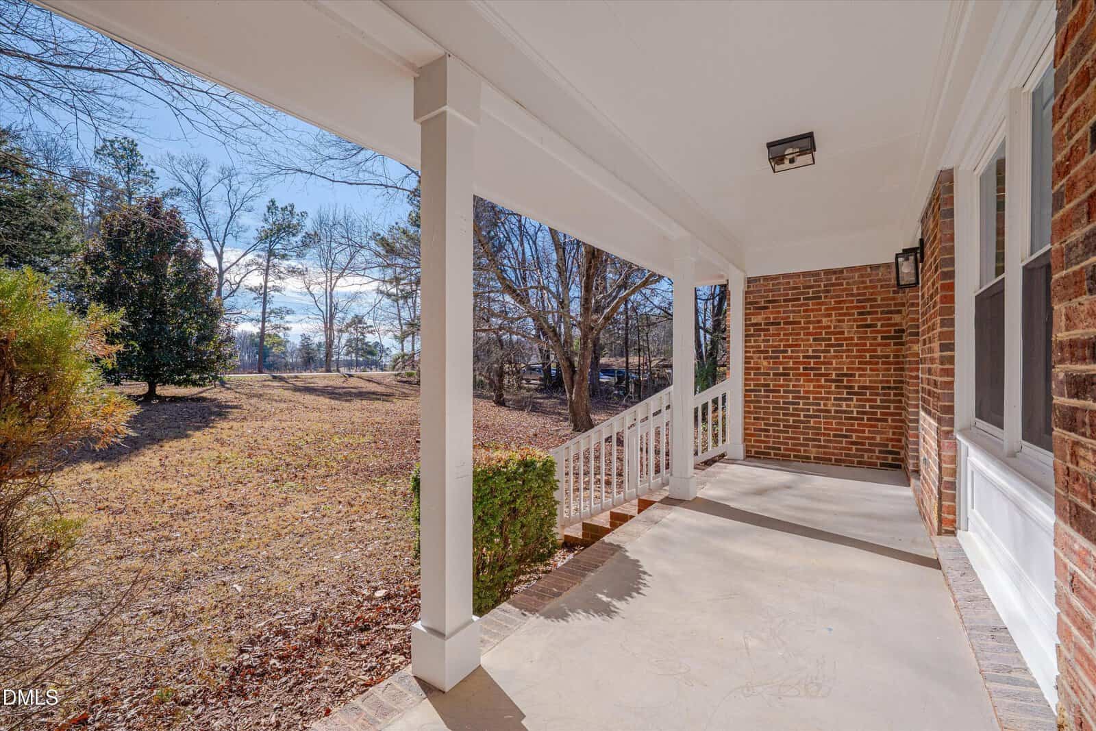 A covered porch with white railings, brick exterior walls, and concrete flooring overlooks a yard with trees and sparse grass under a clear sky.