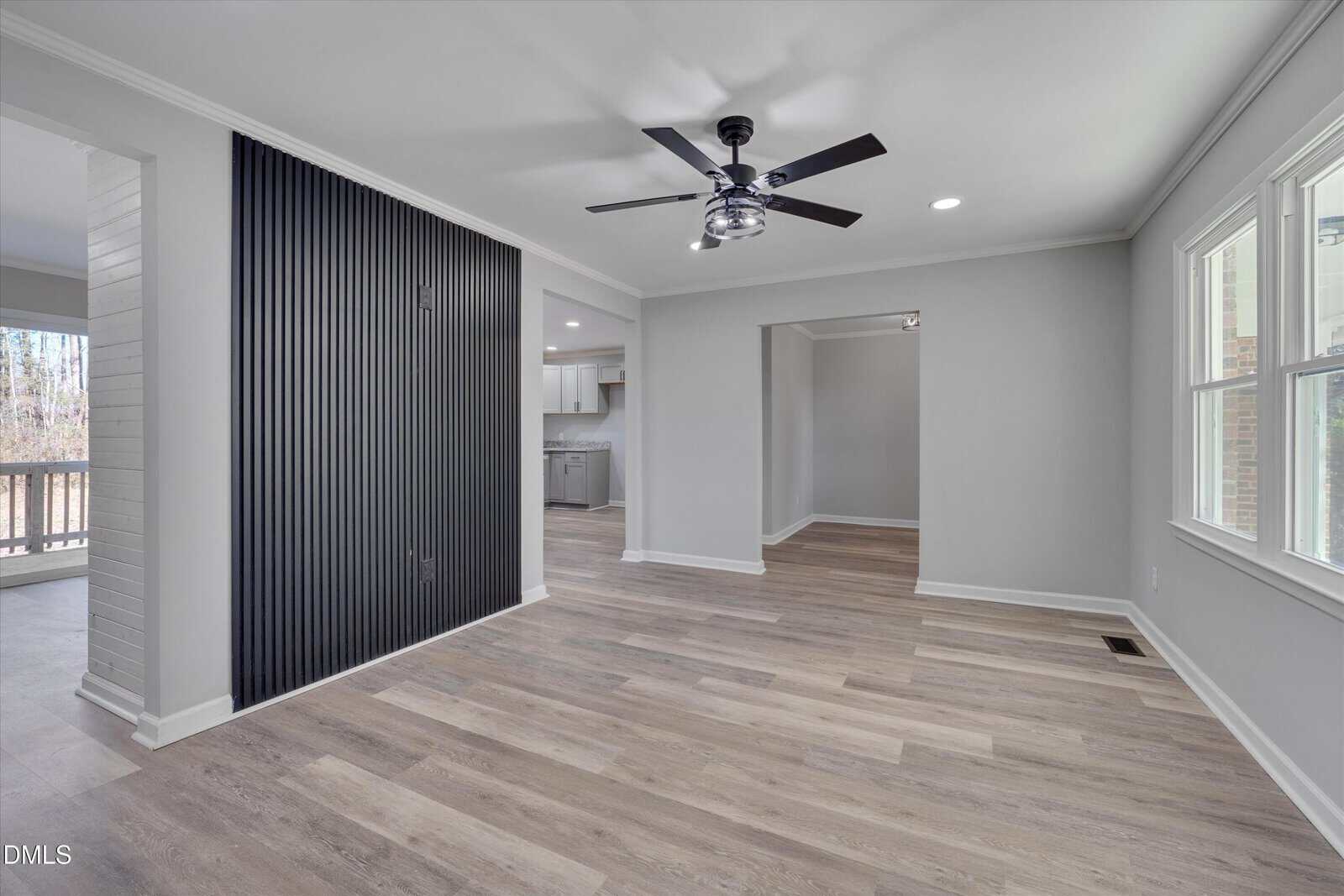 A modern, unfurnished room with light wood flooring, a black ceiling fan, large windows, and a black vertical accent wall. The space leads to a kitchen and another open area, with neutral light gray walls throughout.