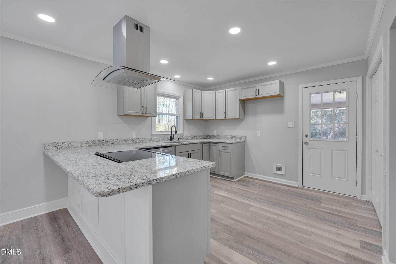 Modern kitchen with light gray cabinets, marble countertops, stainless steel appliances, an island with a stovetop, wood-style flooring, and a door leading outside.