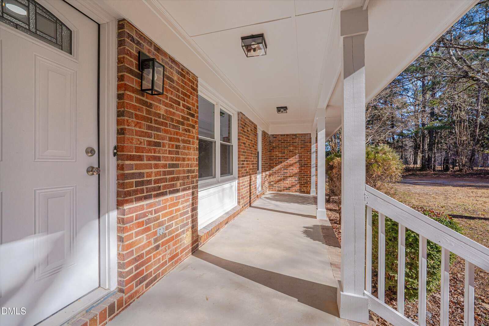 A covered front porch with brick walls, white trim and railing, two windows, two ceiling lights, and a white front door. The porch overlooks a yard with trees and grass.
