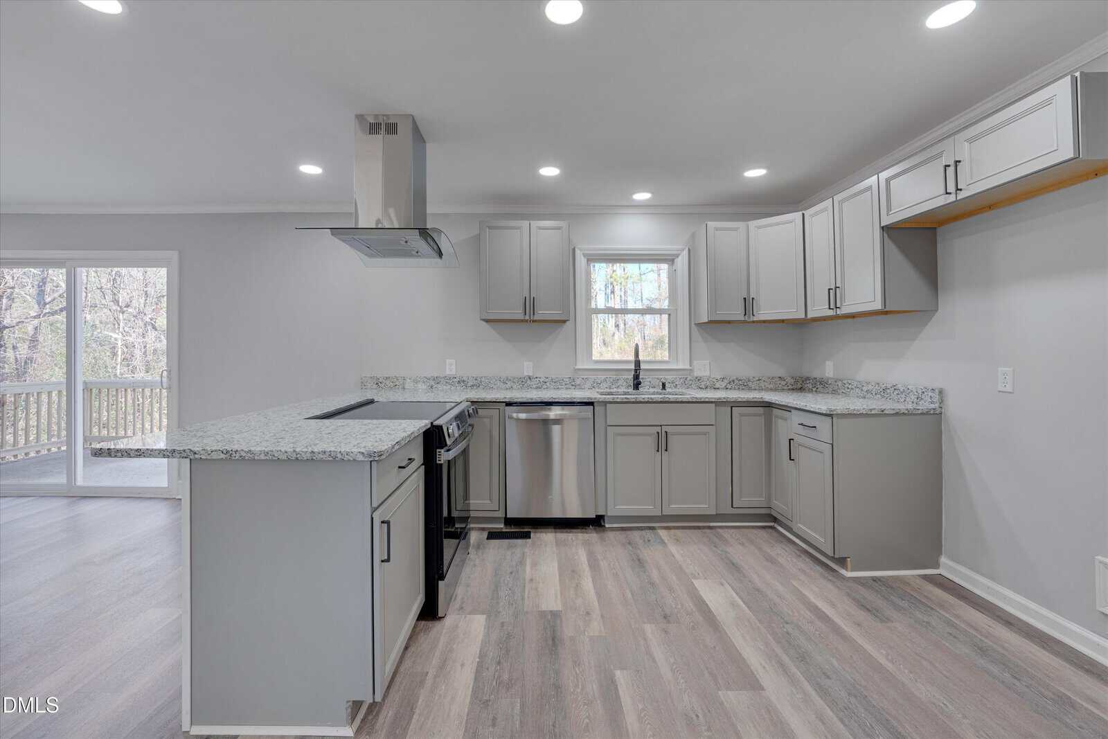 Modern kitchen with light gray cabinets, granite countertops, stainless steel appliances, a center island with cooktop, and wood-look flooring. A window is above the sink, and sliding glass doors open to a deck.