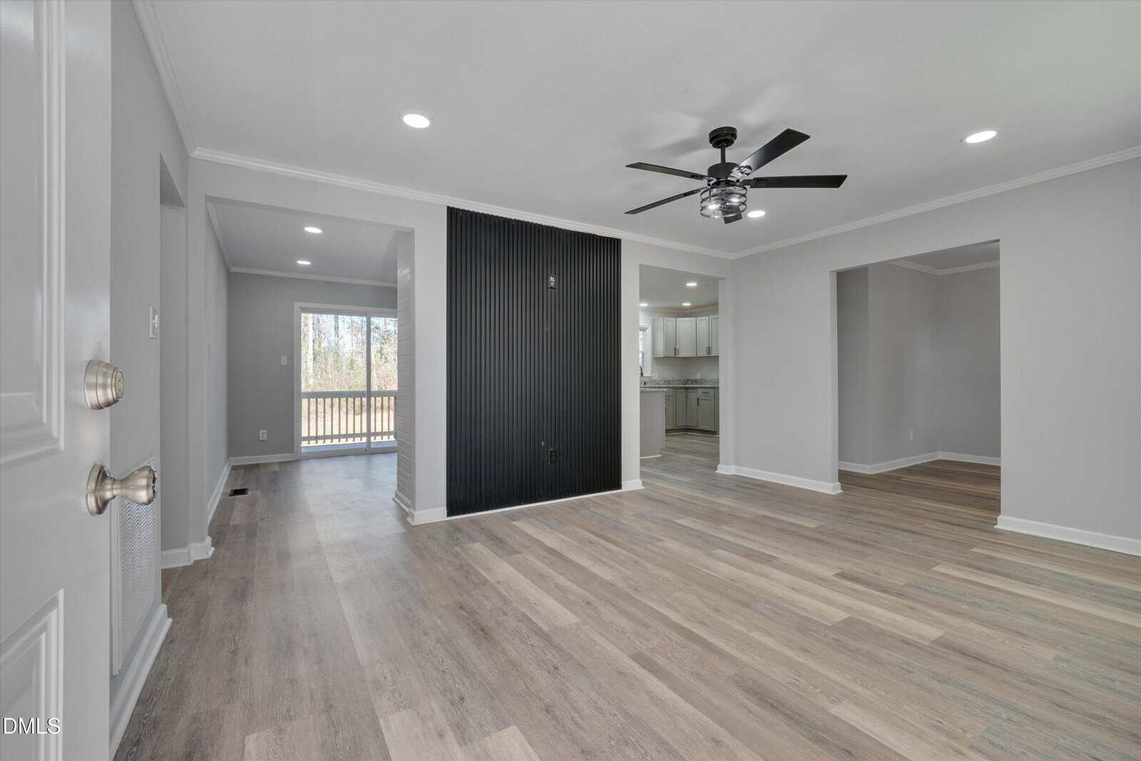 A spacious, modern living area with light wood flooring, a black accent wall with outlets, ceiling fan, and open layout leading to a kitchen and another room. Large window provides natural light at the back.