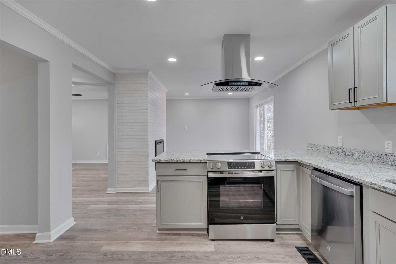 Modern kitchen with light gray cabinets, stainless steel appliances, granite countertops, and wood-style flooring. Open layout leads to a living area with a fireplace and sliding glass doors. Neutral color palette throughout.