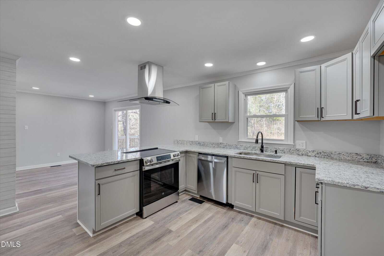 Modern kitchen with light gray cabinets, granite countertops, stainless steel appliances, electric stove with hood, dishwasher, and sink under a window. Light wood flooring and white walls with recessed lighting complete the space.