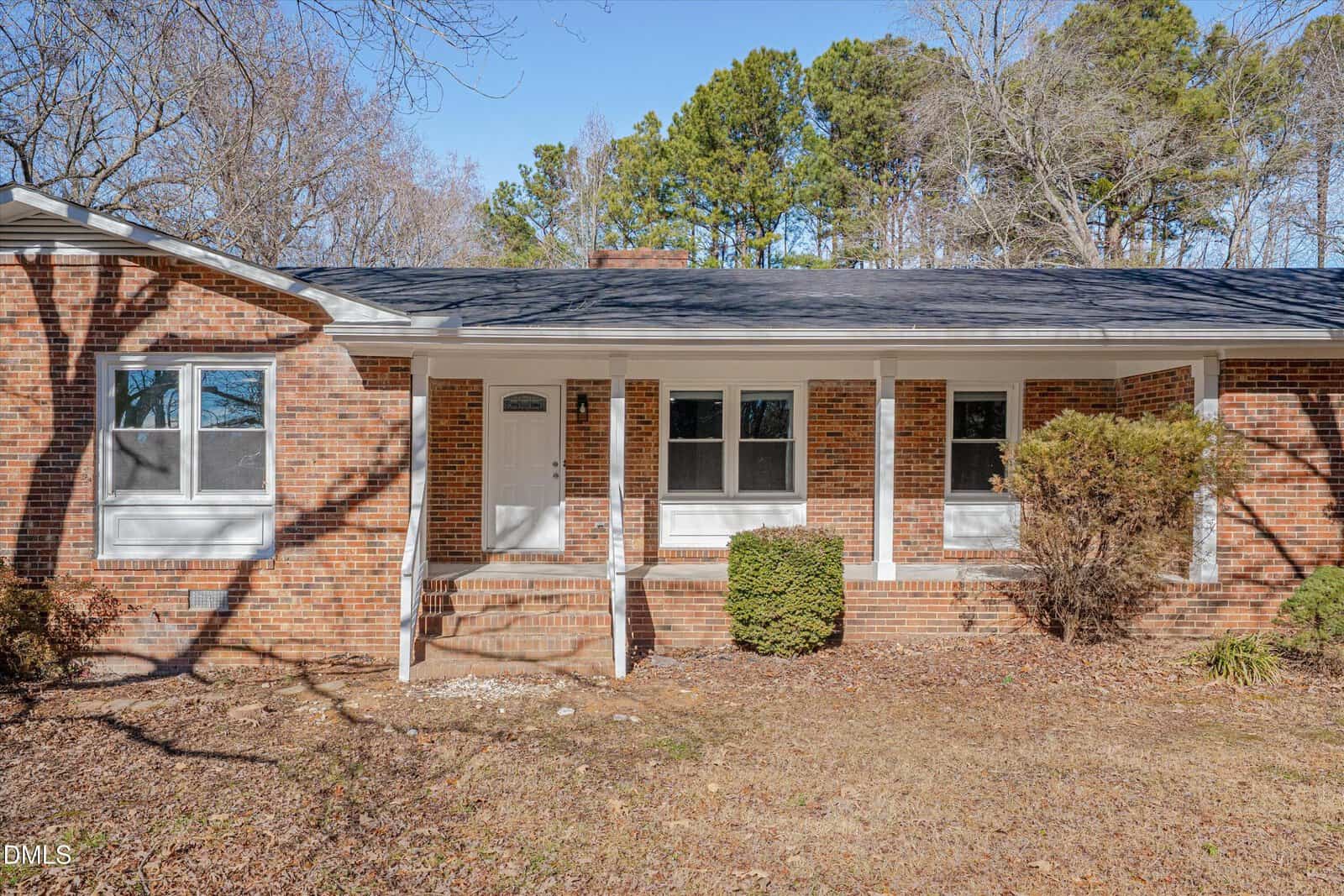 A single-story brick house with a covered front porch, white trim, and three sets of steps. The yard has dry grass, small bushes, and trees in the background under a clear blue sky.