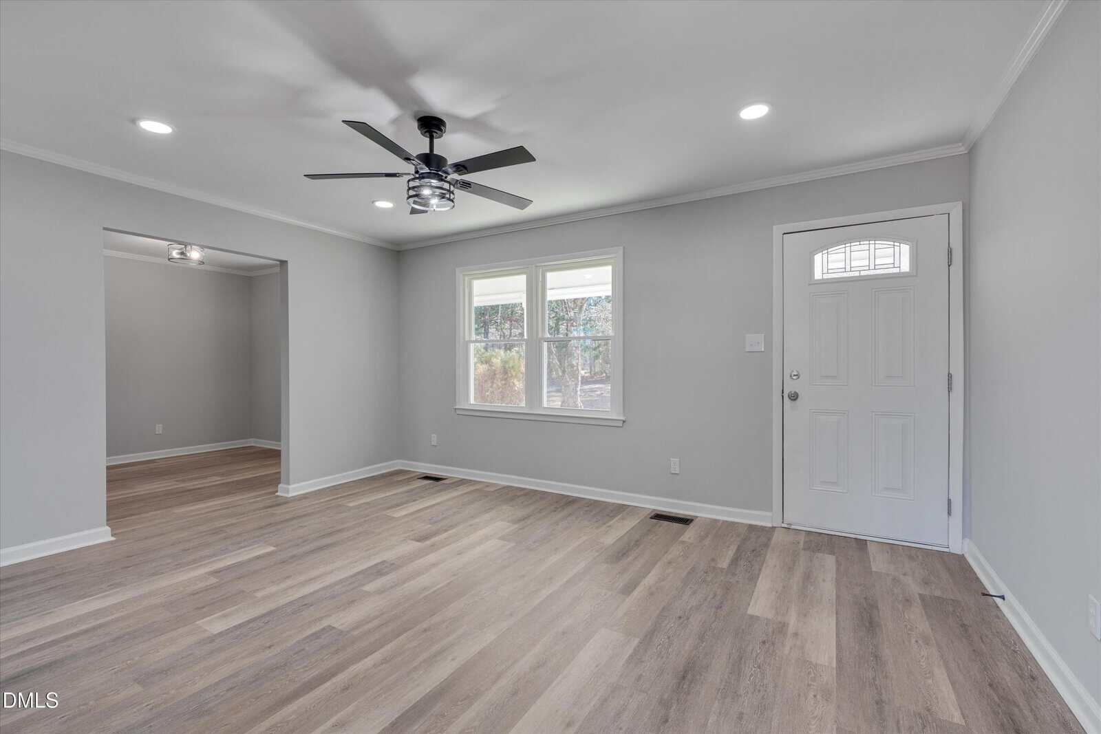 A bright, empty living room with light wood flooring, gray walls, a ceiling fan with lights, two windows, and a white front door. An open doorway leads to another room with similar decor.