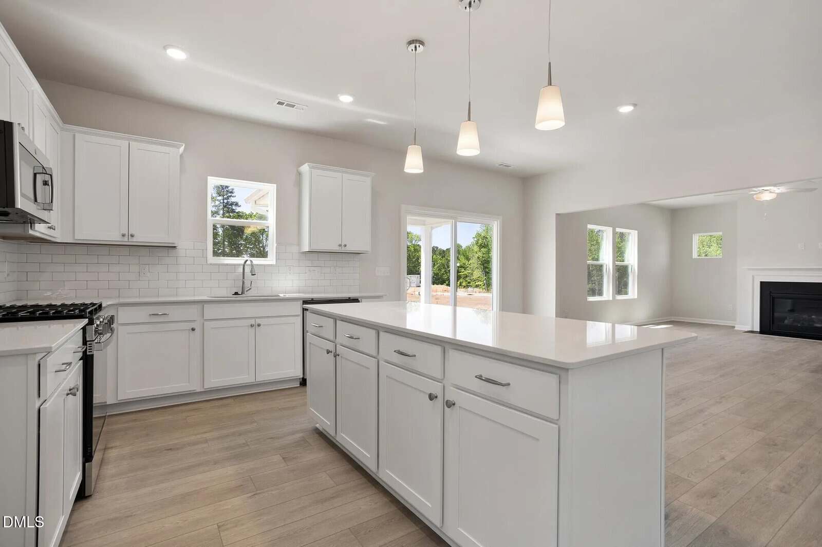 Modern kitchen with white cabinets, stainless steel appliances, subway tile backsplash, large white island, pendant lights, and light wood floors. Open layout leads to a living area with a fireplace and large windows.