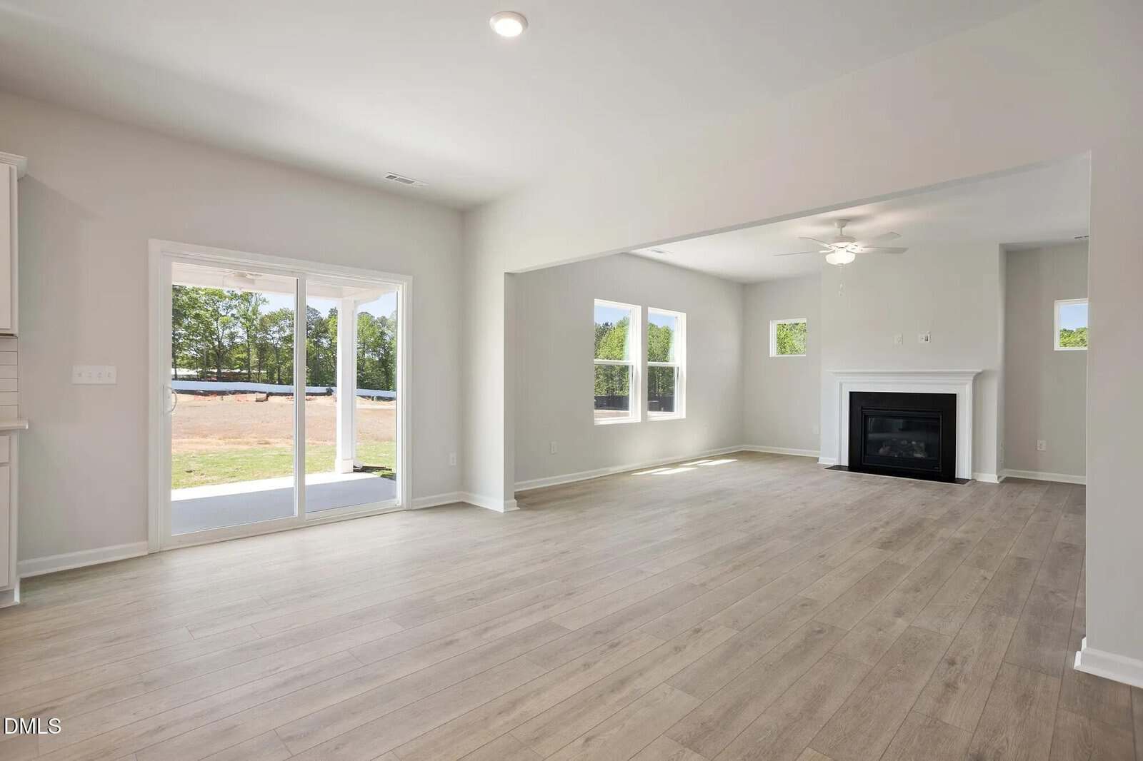 Spacious, empty living room with light wood flooring, large windows, sliding glass door to a patio, and a white fireplace. Walls are painted light gray, and the room is filled with natural light.