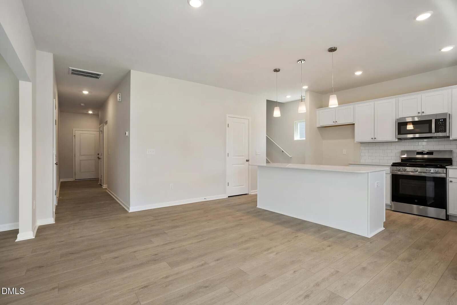 Modern kitchen with white cabinets, stainless steel appliances, a large island, pendant lights, and wood flooring. Open layout connects to a hallway and entrance. Walls are painted light beige.