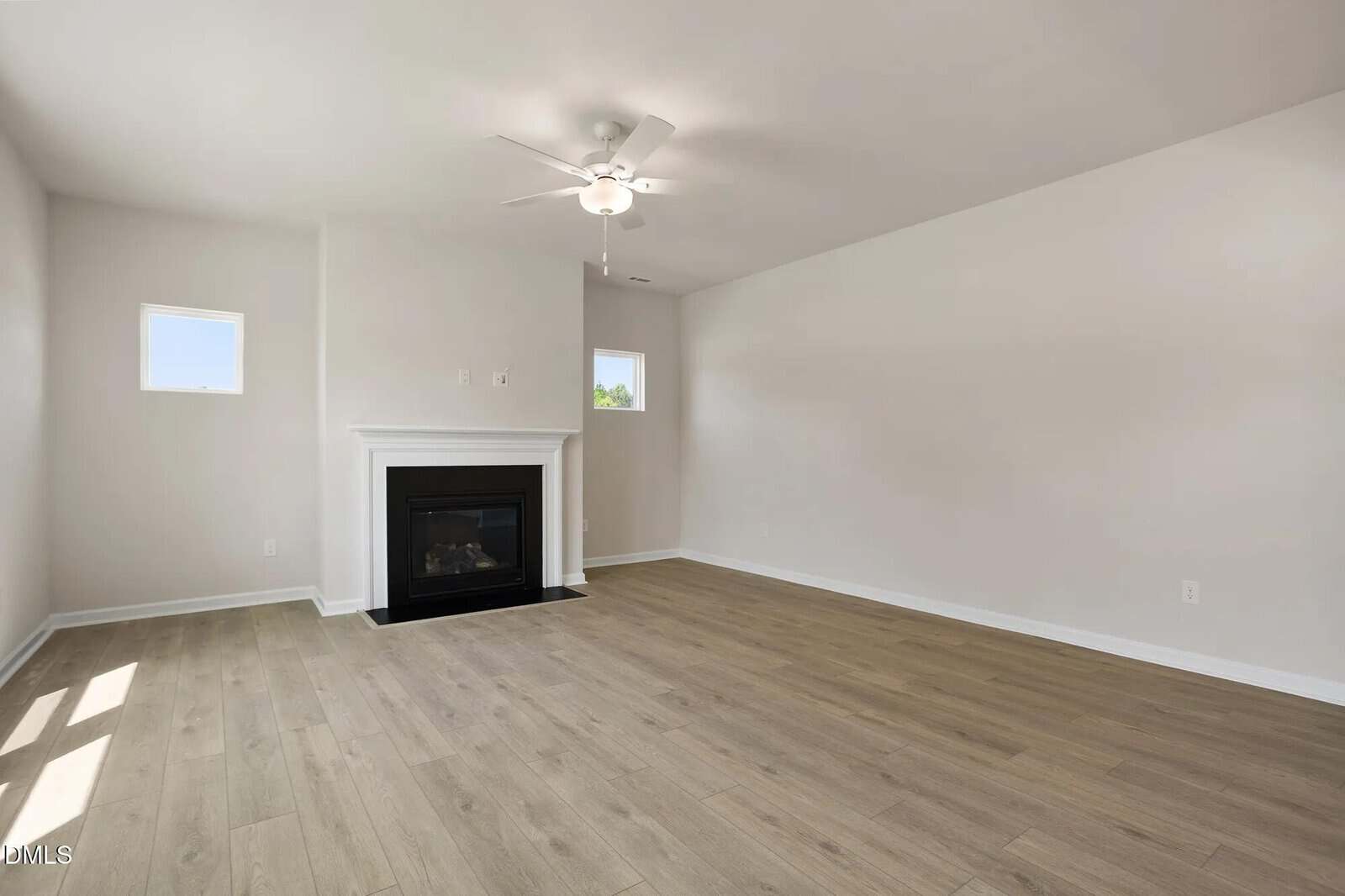 A bright, empty living room with light wood flooring, white walls, two small windows, a ceiling fan, and a black fireplace with a white mantel.
