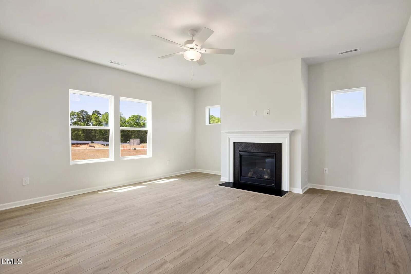 Bright living room with light wood floors, white walls, three windows, a ceiling fan, and a white fireplace with a black insert. Natural light fills the space, highlighting the clean, modern design.