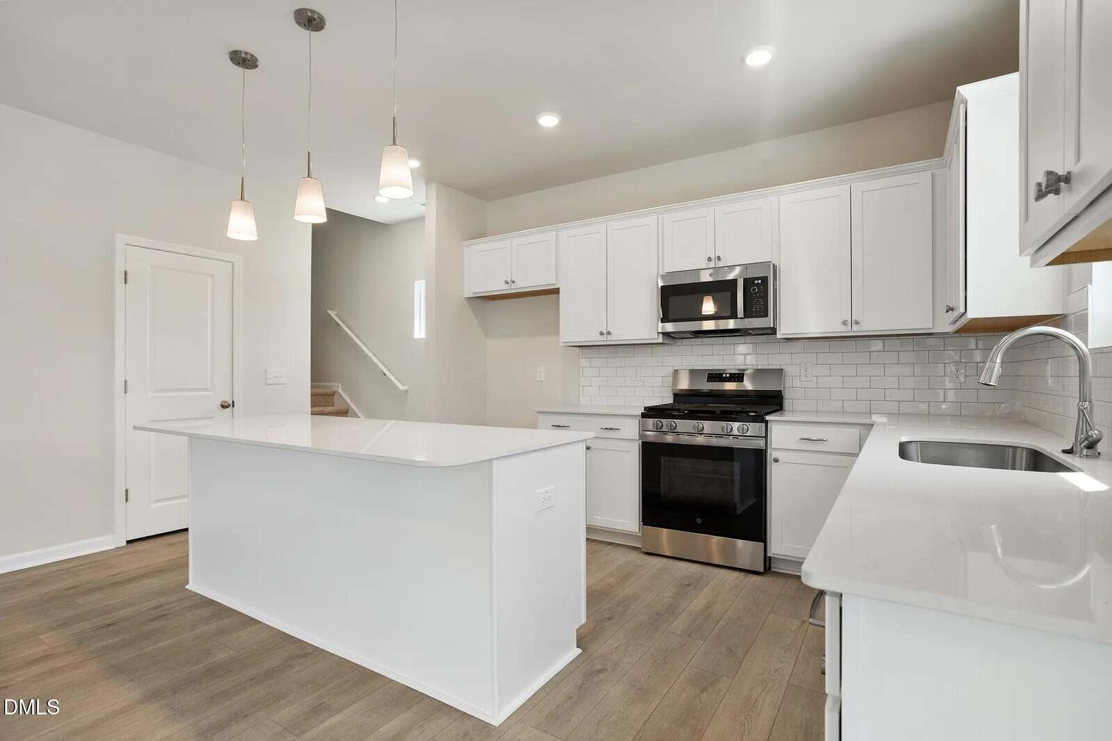 Modern kitchen with white cabinets, stainless steel appliances, subway tile backsplash, a large white island with pendant lights, light wood flooring, and a staircase visible in the background.