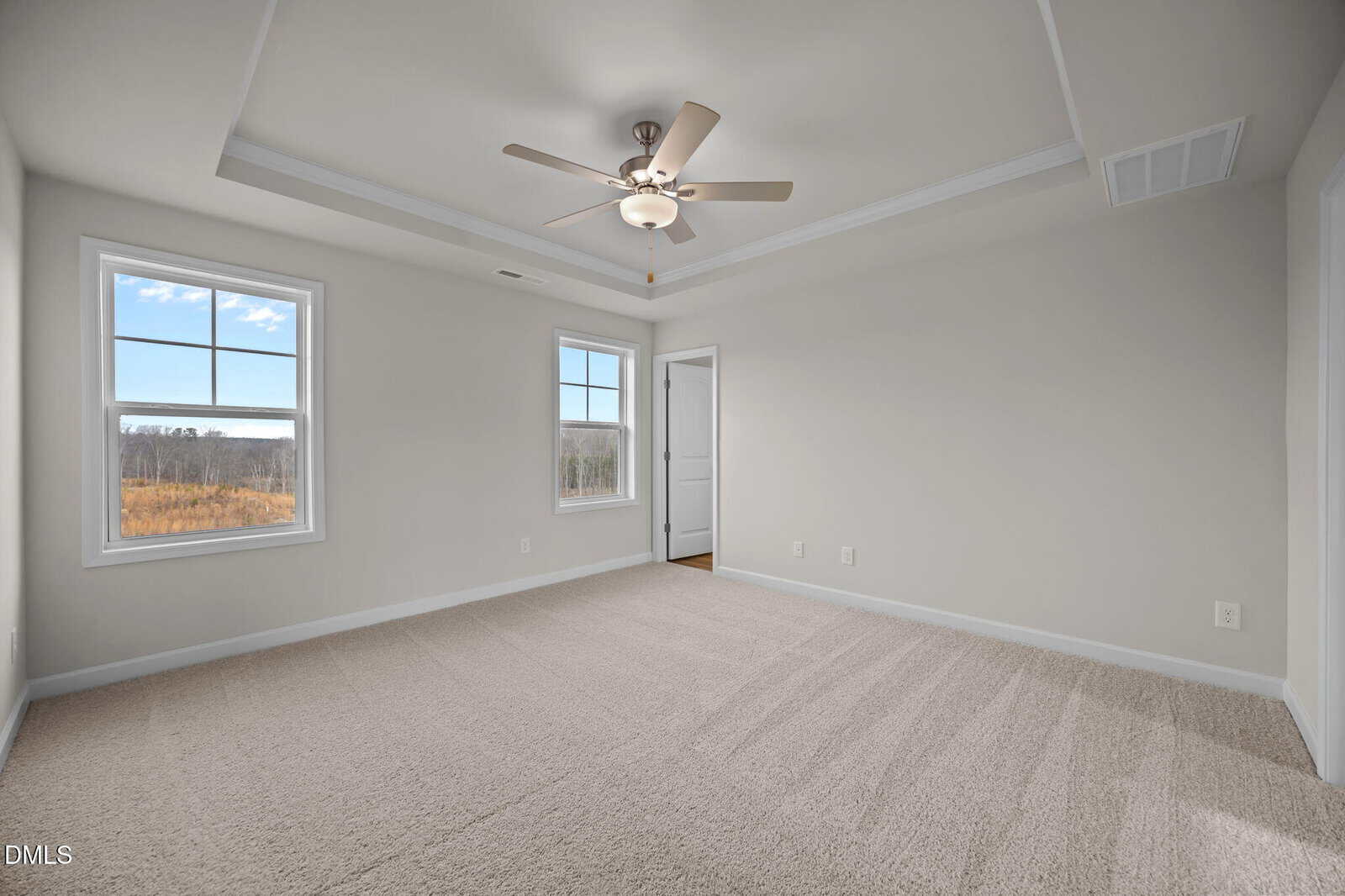 A bright, empty bedroom with beige carpet, light gray walls, two windows showing a scenic outdoor view, a ceiling fan with lights, and a white door leading to another room.