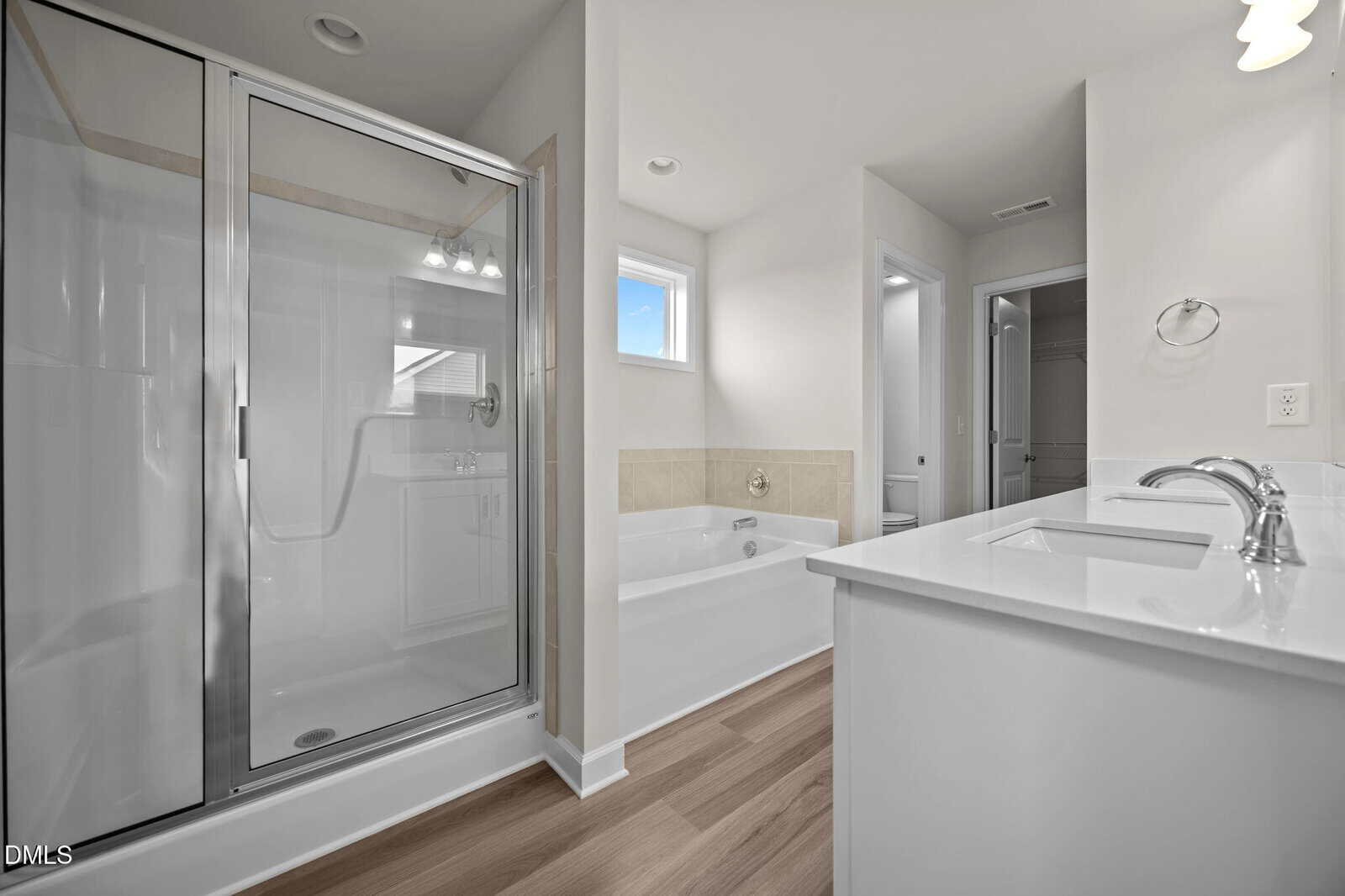 Modern bathroom with glass shower, separate bathtub, white vanity with sink, silver fixtures, light wood flooring, and white walls. A small window lets in natural light near the bathtub.
