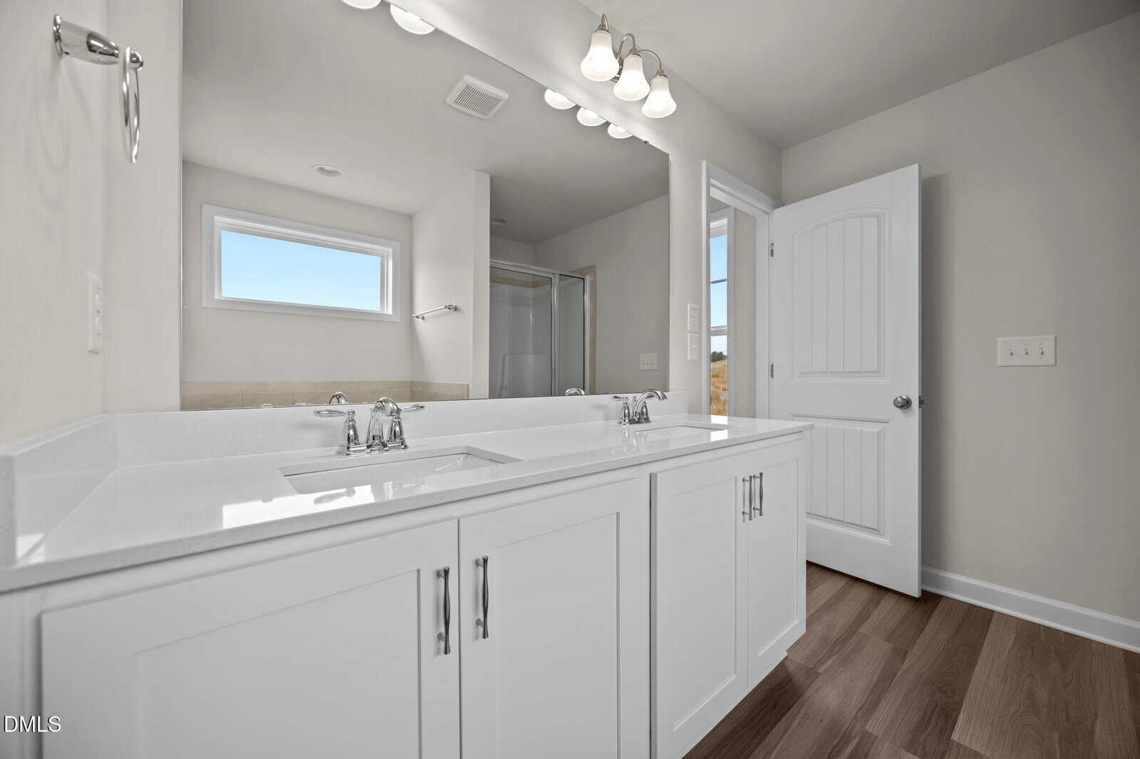 A modern bathroom with double sinks, white cabinets, a large mirror, wood-look flooring, and a shower in the background. Natural light enters through a rectangular window above the sinks.