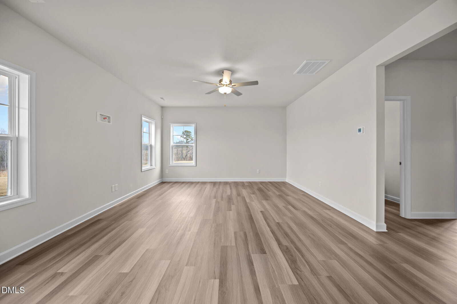 Spacious, empty living room with light wood flooring, white walls, large windows letting in natural light, a ceiling fan with lights, and doorways leading to other rooms.