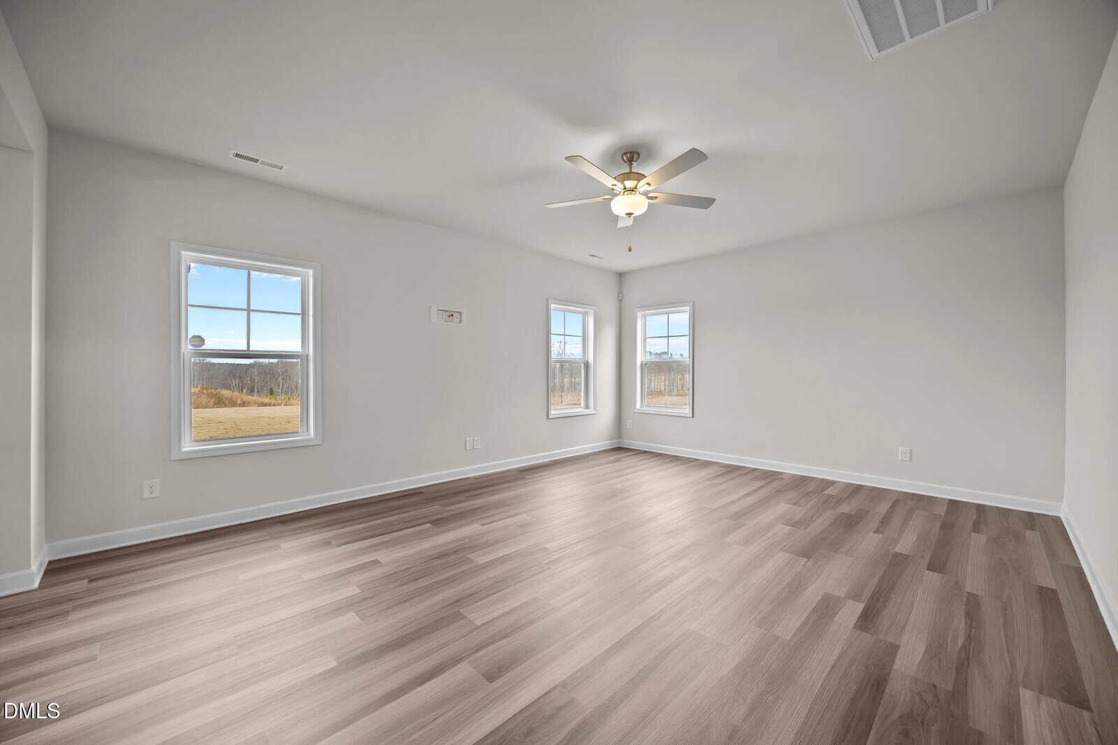 A spacious, empty room with light wood flooring, three windows letting in natural light, white walls, and a ceiling fan with a light fixture.