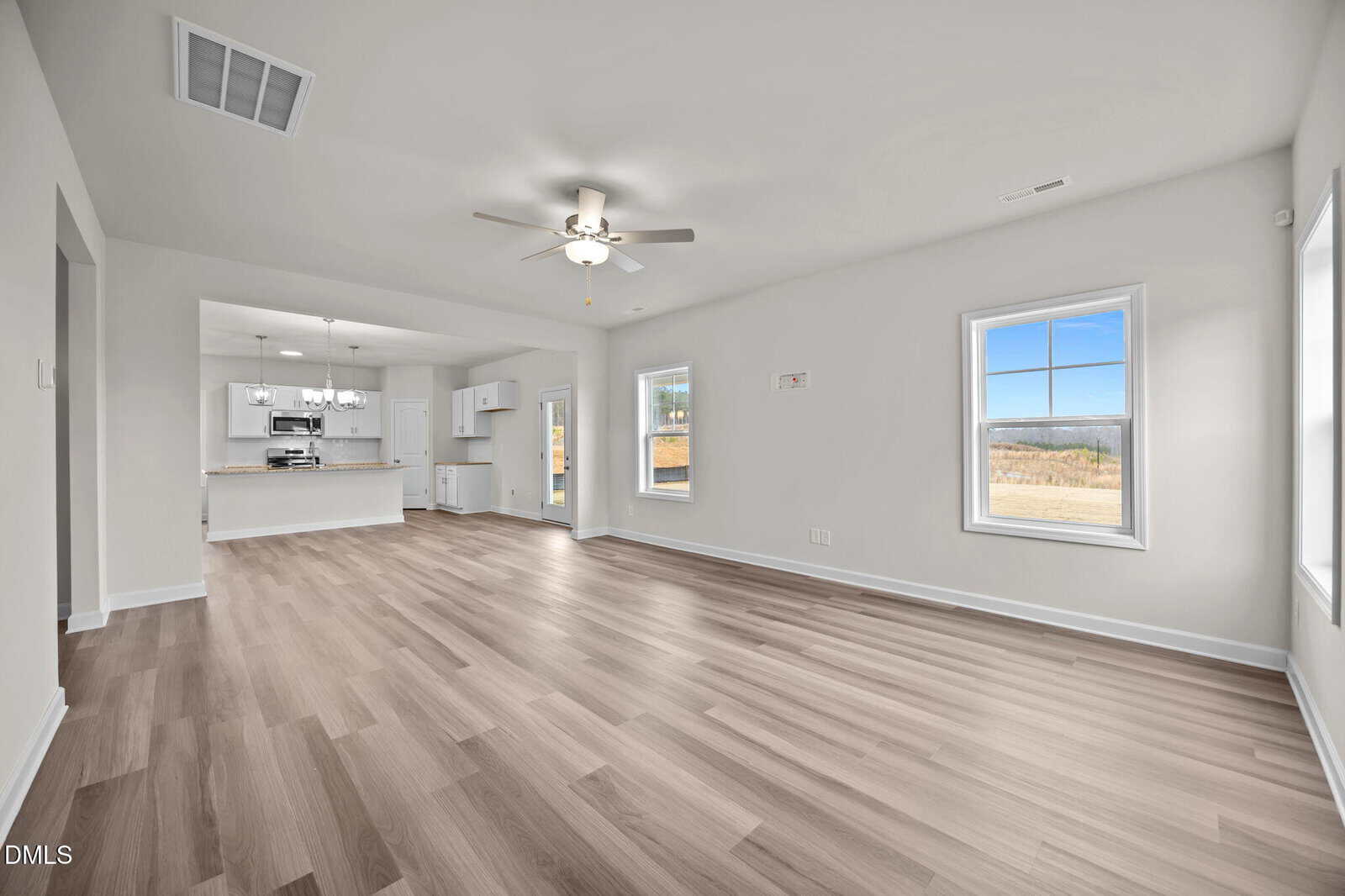 A spacious, bright open-concept living area with light wood flooring, white walls, and large windows. The room flows into a modern kitchen with white cabinets, stainless steel appliances, and a ceiling fan overhead.