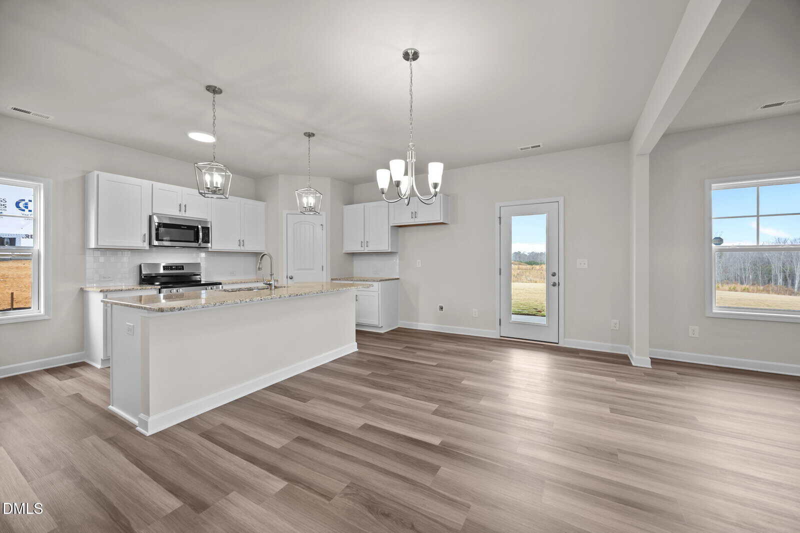 Modern, empty kitchen and dining area with white cabinets, stainless steel appliances, granite countertops, light fixtures, and large windows providing natural light. Light wood flooring extends throughout the open space.
